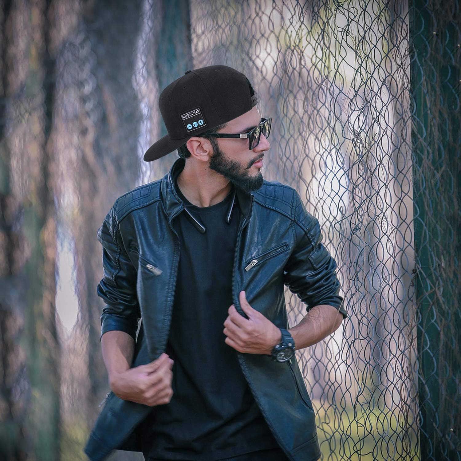 Stylish man in a black leather jacket and baseball cap, posing outdoors against a mesh background.
